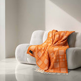 Heating & Plumbing London 'Mosaic Orange' checked wool throw draped over a curved cream sofa in a minimalist, AI-generated modern interior with marble flooring and soft angled sunlight.