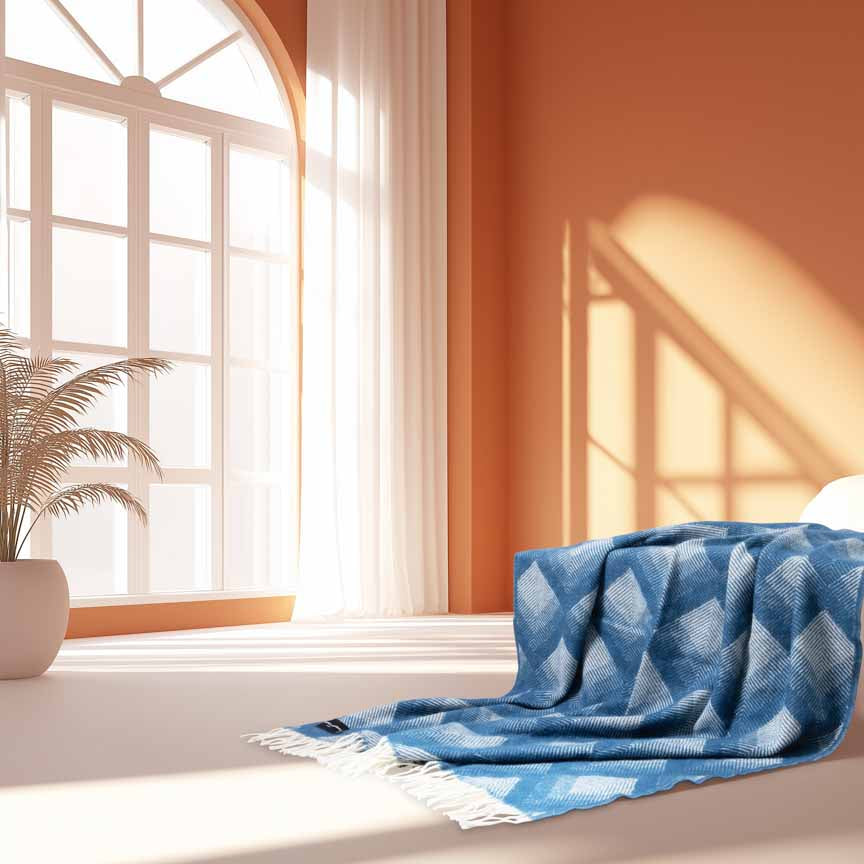 Swinging London wool blanket with white fringe draped across a bed. The product is shown in a sunlit, AI-generated interior with large arched windows, terracotta walls, and a potted palm.