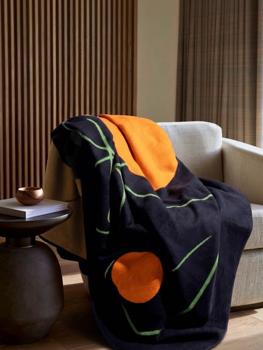 A dark navy throw blanket with abstract orange and green patterns is draped over a light beige armchair in a modern living room setting.