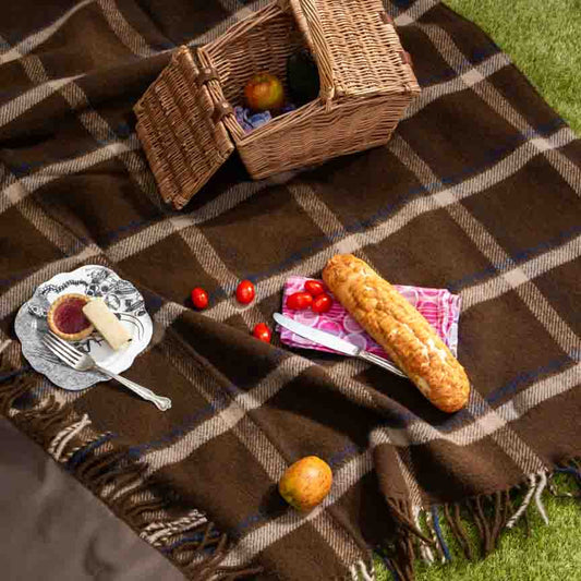  Luxury wool waterproof picnic blanket in brown checkmate pattern, styled with picnic food and wicker basket on grass—perfect for outdoor dining and countryside picnics.